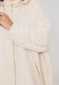 Jasno-beżowa ocieplana kurtka z wysokim kołnierzem i złotym zamkiem, z prążkowanymi mankietami oraz bocznymi kieszeniami z ozdobnymi zamkami.