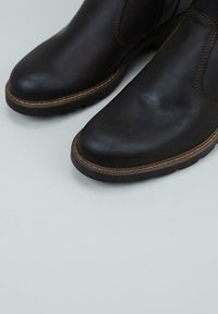 Botas negras de cuero sin cordones con costuras marrones y suelas de goma gruesa sobre un fondo gris claro.