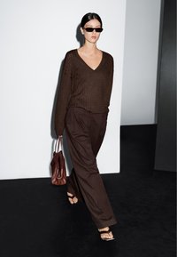 Vrouw die binnen loopt in een bruine V-hals trui, wijde broek, zwarte sandalen, donkere zonnebril en een bruine handtas draagt.