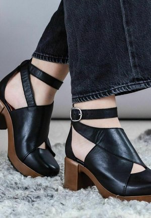 Black leather open-toe shoes with ankle straps, featuring a chunky brown heel. Smooth texture with cut-out design details.