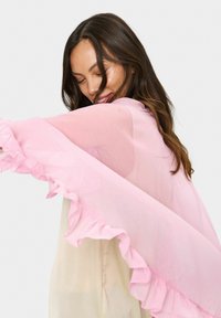 Sheer, light pink cape with ruffled edges. Soft texture, flowing design, layered over a cream-colored garment with a smooth finish.