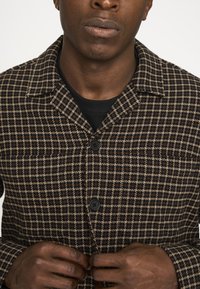 Uldblandet frakke med brun og creme ternet mønster, struktureret krave, sorte knapper og en tekstureret finish. Hænder der justerer knapperne.