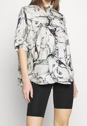 Femme portant une chemise à boutons ample avec un motif abstrait noir et blanc, des manches longueur coude, et un short noir ajusté jusqu’aux genoux.