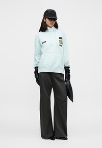 Φούτερ σε μέντα πράσινο με τσέπη μπροστά, μαύρες λεπτομέρειες και άνετη εφαρμογή. Συνδυασμένο με φαρδιά μαύρη παντελόνια και μαύρα δερμάτινα γάντια, κρατώντας τσάντα χειρός.