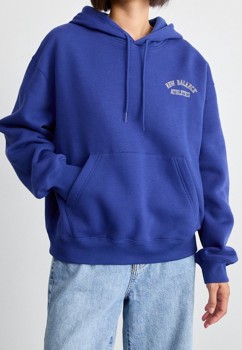 Personne portant un sweat à capuche bleu New Balance Athletics avec poche avant et un jean bleu clair sur un fond uni.