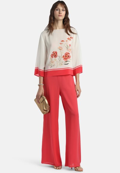 Femme portant un chemisier blanc ample avec des imprimés floraux rouges et un pantalon large rouge, tenant une pochette dorée et portant des sandales.