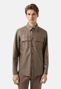 Brun læderskjorte med krave, to brystlommer og knaplukning foran. Glat tekstur og figursyet design. Lange ærmer.