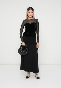 Zwarte fluwelen jurk met een doorschijnende top met lange mouwen, slim fit en een vloeiende rok. Gecombineerd met een zwarte handtas en platte schoenen.