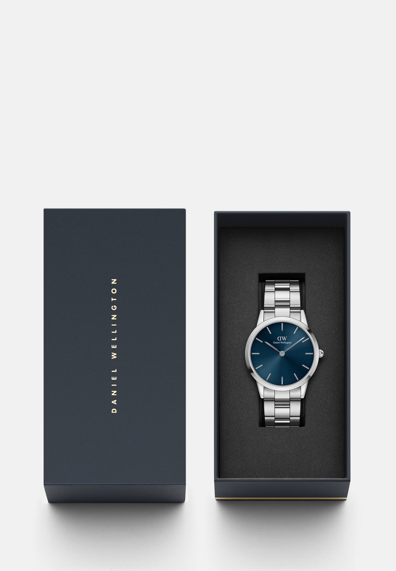 Montre en métal argenté avec un cadran bleu, présentant un design minimaliste, placée dans une boîte noire avec un logo doré et un rembourrage intérieur doux.
