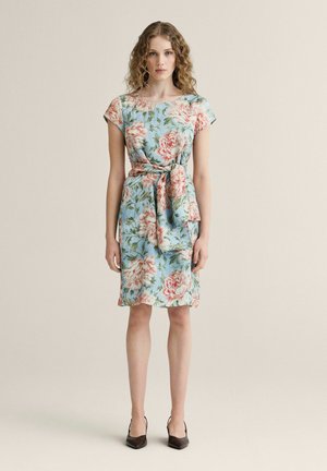 Kvinde med krøllet hår iført en lyseblå blomsterkjole til knæet med bånd i taljen, stående i sorte spidse hæle foran en ensfarvet baggrund.