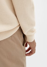 Pull beige texturé avec poignets côtelés, associé à un pantalon marron clair ; main reposant dans la poche, mettant en valeur une coupe décontractée et confortable.