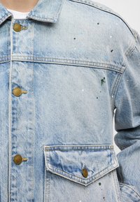 Ανοιχτό μπλε τζιν μπουφάν με κουμπιά στο μπροστινό μέρος, δύο τσέπες στο στήθος και πολύχρωμες πιτσιλιές μπογιάς. Διαθέτει μεταλλικά κουμπιά και κλασικό σχέδιο.