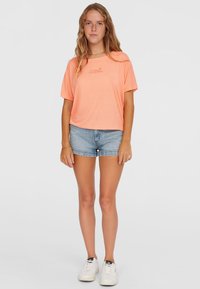 Hellorange T-Shirt mit kurzen Ärmeln und rundem Halsausschnitt, bestickt mit einem Logo, kombiniert mit Jeansshorts und weißen Sneakers.