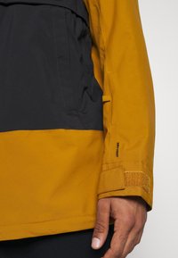 Gros plan sur la main d'une personne et la manche de sa veste en jaune moutarde et noir avec une sangle Velcro au poignet et le logo "DRYVENT".