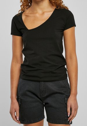 Kobieta ubrana w czarny T-shirt z krótkim rękawem i dekoltem w serek oraz ciemnoszare szorty jeansowe z bocznymi kieszeniami, stojąca na jasnym tle.