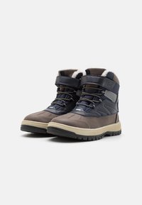 Par de botas de invierno gris y azul marino con suelas negras, cordones y correas de velcro, forradas con lana blanca en el interior.
