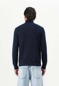 Bărbatul stă cu spatele, purtând un pulover din tricot cu nervuri de un albastru închis și blugi din denim de un albastru deschis, cu buzunarele din spate vizibile.