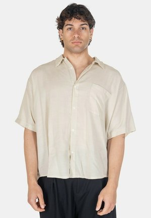 Uomo con capelli ricci indossa una camicia beige a maniche corte e bottoni, ampia, e pantaloni neri, in piedi su uno sfondo chiaro uniforme.