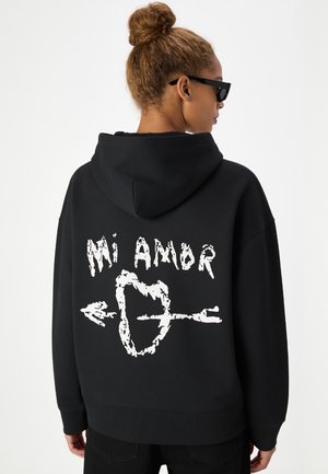 Personne aux cheveux châtain clair en chignon porte des lunettes de soleil noires et un sweat à capuche noir avec un texte blanc "Mi Amor" et un cœur transpercé par une flèche dans le dos.