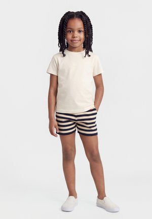 Noor tüdruk seisab käega taskus, kandes kreemikat t-särki, tumeliste ja kreemjate triibuliste lühikeste pükste ning valgeid libisevaid jalatseid valge tausta ees.