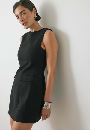 Black dress made of textured fabric, sleeveless with a fitted silhouette, two front pockets, and a unique silver cuff bracelet on the wrist.