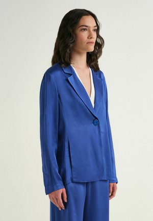 Femme aux cheveux bruns ondulés, portant un blazer bleu roi et un pantalon assorti, debout de profil sur un fond clair uni.