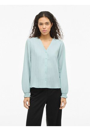 Šviesiai mėlyna plisuota palaidinė su V-formos iškirpte, sagomis priekyje ir ilgomis rankovėmis su surinktais rankogaliais. Derinama su juodomis kelnėmis.