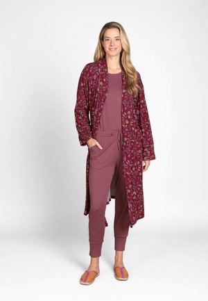 Woman with long blonde hair wearing a maroon striped jumpsuit, floral patterned robe, and orange slip-on sandals, standing on a white background.