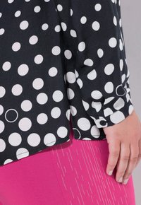 Blusa nera con pois bianchi, caratterizzata da polsini con bottoni e una vestibilità ampia. Abbinata a un indumento rosa con sottili linee verticali.