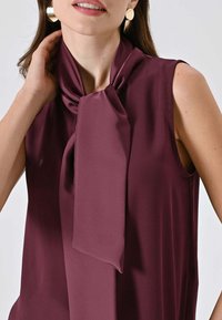 Blusa senza maniche color bordeaux con dettaglio di nodo al collo, realizzata in tessuto liscio. Presenta una vestibilità ampia e una leggera lucentezza. Orecchini rotondi dorati visibili.