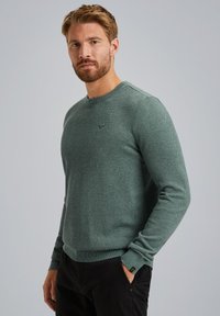 Pull en tricot vert avec un col rond, un motif texturé et des poignets côtelés. Présente un logo discret sur la poitrine.
