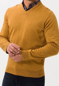 Pull en maille jaune moutarde avec col en V, manches longues et poignets ainsi que bas côtelés. Détail subtil du logo sur la poitrine.