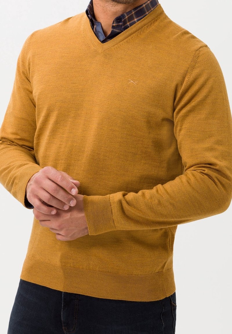 Pull en maille jaune moutarde avec col en V, manches longues et poignets ainsi que bas côtelés. Détail subtil du logo sur la poitrine.