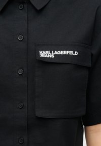 Črna srajca s gumbi z razporkom na prsnem žepu, na katerem je belo vezen napis "KARL LAGERFELD JEANS".