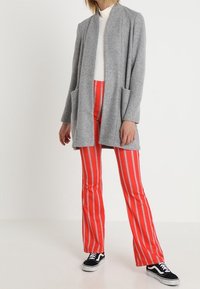 Světlounce šedý kardigan s dlouhými rukávy, otevřeným předním dílem a dvěma bočními kapsami, v kombinaci s červenými rozšířenými kalhotami s proužky a černými teniskami.