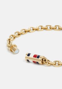 Kuldne käevõru, millel on tsilindriline lukustus, millel on tumesinised, valged ja punased triibud ning graveeritud tekst "HILFIGER".