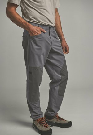 Mann trägt graue Hose mit verstärkten Kniepolstern, beige Wanderschuhe mit orangen Schnürsenkeln und ein hellgraues kurzärmliges Hemd.