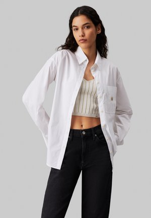 Camicia bianca con colletto a punta, maniche lunghe e taschino sul petto, indossata sopra un top a canotta a coste beige chiaro e jeans neri.