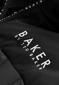 Crni materijal s tekstom "BAKER by TED BAKER" u bijeloj boji. Tekstura je glatka, s blagim sjajem. Detalji sa zatvaračem su vidljivi duž rubova.