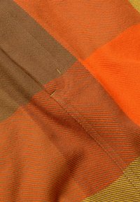 Detailní záběr texturované látky s protínajícími se pruhy v oranžové, hnědé a žluté barvě a viditelnými švy.