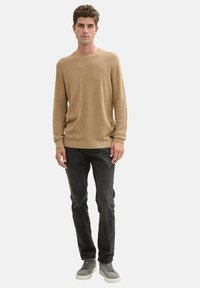 Beige gestrickter Pullover mit Rundhalsausschnitt und langen Ärmeln, kombiniert mit dunklen Jeans und hellgrauen Turnschuhen. Strukturiertes Stoffdetail sichtbar.