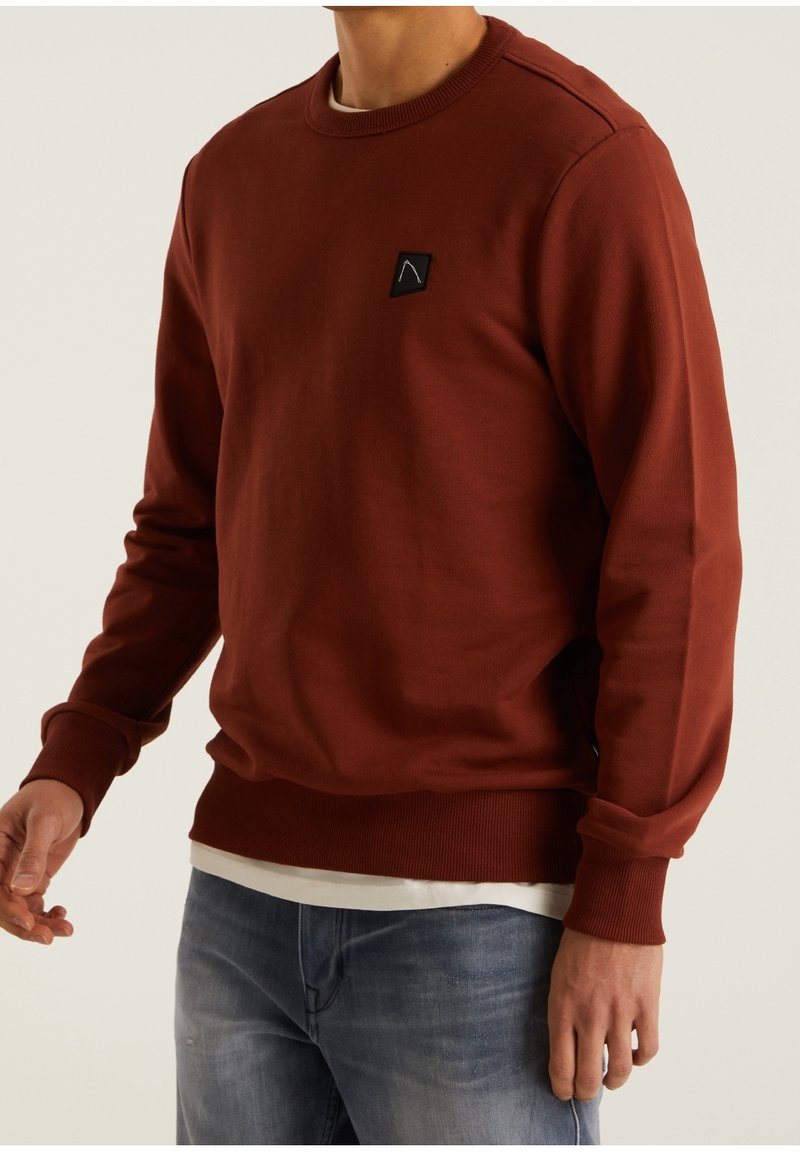 Homme portant un sweat-shirt ras du cou de couleur rouille avec un petit logo noir sur la poitrine, superposé à une chemise blanche et assorti d'un jean bleu.