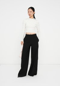 Weißes langärmliges Cropped-Top mit texturiertem Muster, kombiniert mit hochtaillierten schwarzen weiten Hosen mit seitlichen Taschen und einem lässigen Schnitt.