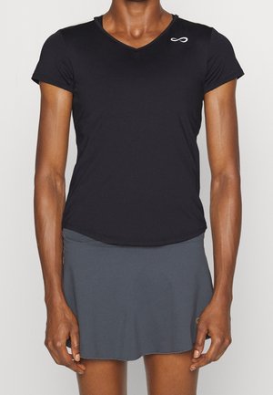 Person trägt ein schwarzes kurzärmeliges V-Ausschnitt-Oberteil mit kleinem weißem Logo und einen grauen Sportrock vor einem einfarbigen Hintergrund.