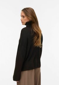 Ciemnobrązowy dziany sweter z wysokim kołnierzem, opuszczonymi ramionami i prążkowanymi krawędziami. Noszony z szerokimi spodniami, pokazującymi luźne dopasowanie.