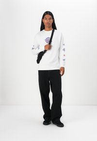White long-sleeve t-shirt with graphic prints on sleeves, paired with black wide-leg pants and black sneakers. Black crossbody bag is worn.