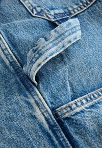 Tessuto in denim blu con una superficie texture, caratterizzato da una tracolla piegata e cuciture visibili lungo i bordi e le tasche.