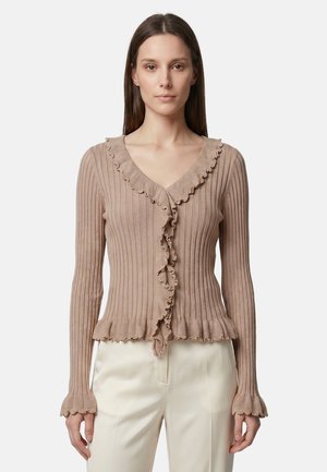 Femme portant un cardigan beige côtelé avec des bords à volants et un pantalon crème, debout devant un fond blanc uni.