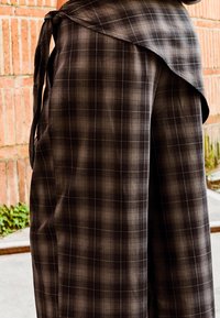 Oseba, oblečena v črno-sivo karo ovijalno krilo, stoji na prostem poleg opečne stene in betonske talne površine.