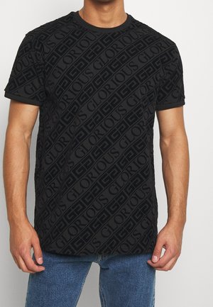 Uomo che indossa una T-shirt nera a maniche corte con un motivo diagonale ripetuto della parola "GLORIOUS" e forme geometriche, abbinata a jeans blu.